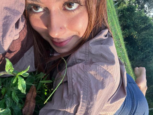 DamiAmbani with brown eyes, smiling, wearing a pinkish shirt, lying in grass, holding green plants, her bare ass slightly visible, sunlight on her face.