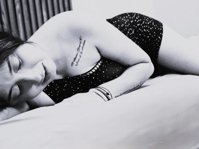 JuliethCruz with her eyes closed, lying on a bed in black polka dot underwear, showing a tattoo on her right shoulder and another on her left forearm.