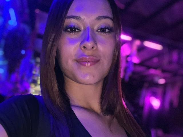 DamiAmbani with glossy lips, brown hair, black top, nose ring, and purple-blue lighting, smiling slightly. Her ass is not visible. She has a cute, sexy look.