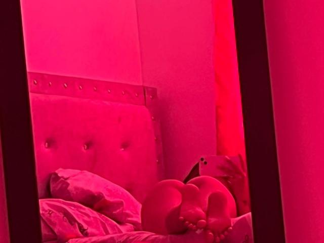 Red-lit mirror shot of DamiAmbani's ass and feet, lying on a bed with a tufted headboard and pink pillows, surrounded by red lighting.