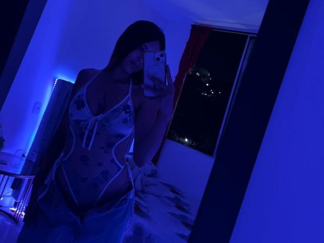 DamiAmbani in a sheer, floral lace-up bodysuit, showing her tits and pussy through the fabric, taking a mirror selfie in blue-lit bedroom, holding a phone.