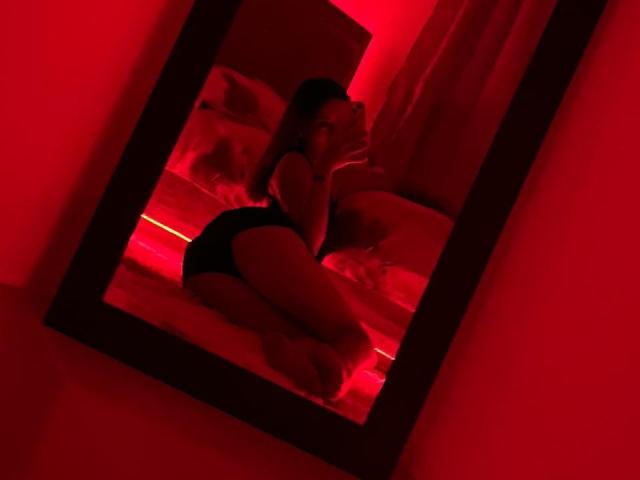 Red-lit mirror showing DamiAmbani kneeling on bed, wearing black panties, back to camera, with her ass up and her hand near her mouth, looking over shoulder.