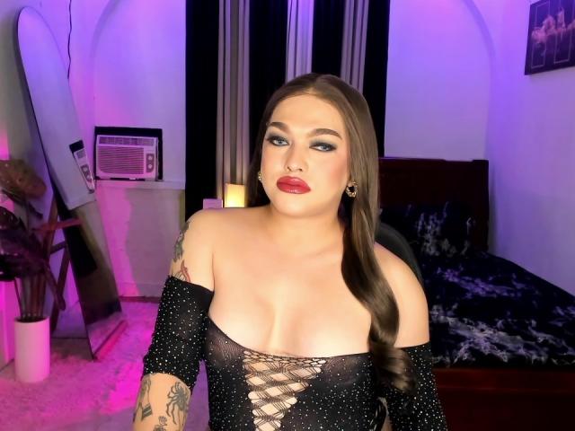 BoobsieBigCock with long brown hair, blue eye makeup, red lipstick, black off-shoulder top with lace, showing tits, tattoo on left arm, standing in room with purple lighting.