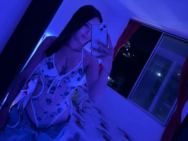 DamiAmbani with blue-lit room, wearing a floral, cut-out, white lingerie top, holding a phone, mirror selfie, long black hair, tattoos visible, red curtains, window in background.