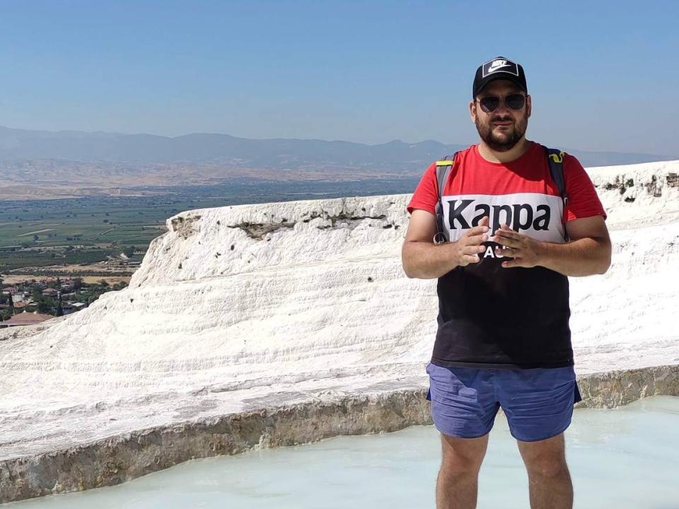 DamienBlack, een wat dikkere man met een baard en zonnebril, gekleed in een rood en zwart shirt, blauwe korte broek en een pet, staat voor een witte rots en houdt een camera vast..