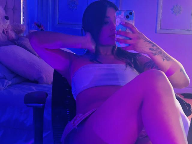 KatianPierce mit dunklem Haar, einem weißen Top und pinken Shorts, macht ein Selfie vor einem Spiegel mit violetter und blauer Beleuchtung, die Arme hinter dem Kopf, zeigt Tattoos und hält ein Handy..