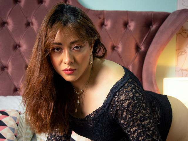 DannaRiva with wavy brown hair, wearing a black lace top, leaning on a bed, showing her tits, looking at the camera with a sultry expression.
