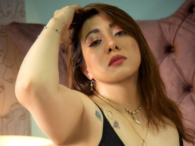 DannaRiva with brown hair, looking up, arm raised, wearing black lace top, gold necklace, earrings, small tattoos on arm and chest, slightly parted lips, pinkish background.