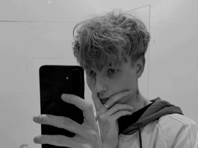 Aaron, con i capelli biondi spettinati, che tiene un telefono, coprendosi la bocca con la mano, indossa una felpa, ha un anello al dito, sta guardando un selfie allo specchio, foto in bianco e nero, viso leggermente sporco..