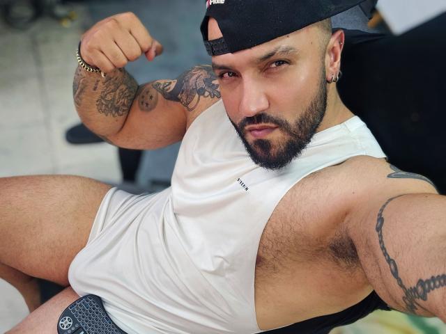 BardoCkroy69, muscular, bearded, wearing a white tank top, flexing bicep, tattoos on arm and chest, black cap, gray shorts, sitting, looking at camera, slightly hairy chest.
