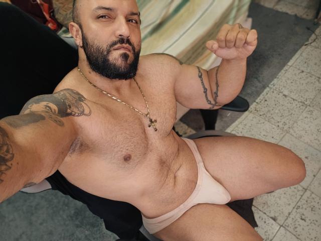 BardoCkroy69, a muscular, bearded man with tattoos, wearing pale pink briefs, sitting, showing a fist, looking at the camera, with a cross necklace.