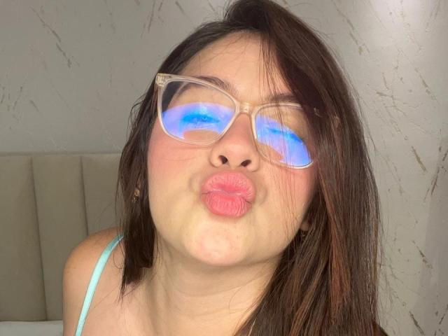 CaroliineRoss has shoulder-length brown hair, wearing clear, oversized glasses with blue reflective light on the lenses. She's puckering her lips in a kissy face. She has fair skin and is wearing a light blue strap, possibly from a top. Her hair is slightly messy, and her expression is playful. The background is a plain, textured wall. The image is a close-up, focusing on her face.