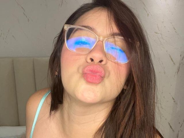 CaroliineRoss with blue glowing glasses, puffy pink lips, and straight brown hair, making a puckered face. She's wearing a light blue strap.