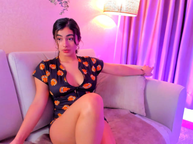 Emma Beaumont, com cabelo escuro, vestindo um vestido preto com estampas laranja, sentada em um sofá branco, com as pernas cruzadas, mostrando os seios, com o braço apoiado no sofá, sob uma luz roxa e rosa..