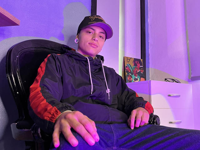 Wailie, portant une veste noire avec des rayures rouges, un jean bleu et une casquette noire, est assis sur une chaise, regardant la caméra avec un léger sourire, et a des piercings aux oreilles..
