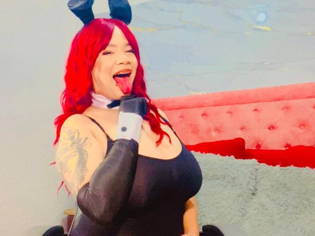 VictoriaField with red hair, black bunny ears, and a black bodysuit, sticking her tongue out, showing her big tits, and a tattoo on her right arm.