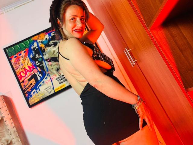DolcceFerrer with a smile, back to camera, black dress with sideboob, black hair in a ponytail, hand on her ass, wooden door, red lighting, bracelet on wrist.