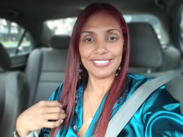 Fuego69X with red hair, smiling, wearing a blue shiny shirt, seatbelt, and earrings, sitting in a car, holding shirt collar with right hand.