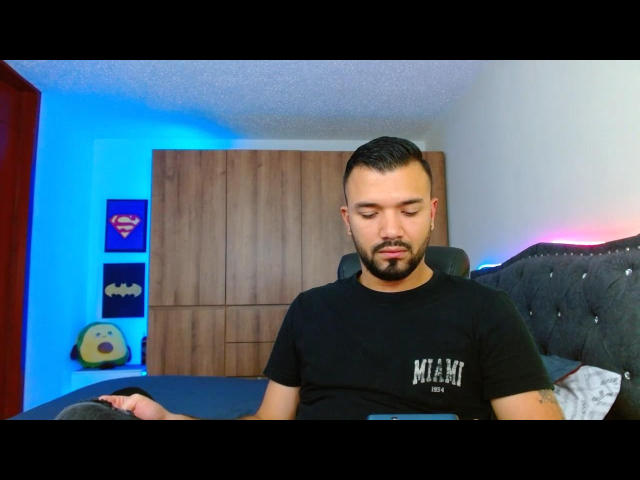 MathewwHot, a bearded dude with short black hair, wearing a black t-shirt, sitting on a bed, eyes closed, looking relaxed, with a tattoo on his right arm.