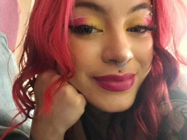 DahianaKlimt with red hair, yellow and pink eyeshadow, long eyelashes, blue nose ring, purple lipstick, and a nose piercing, smiling with hand on cheek.