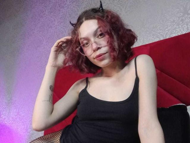 SophieBlake with curly red hair, small black horns, glasses, and a black tank top, showing her tits, sitting on a red cushion, holding her hair.