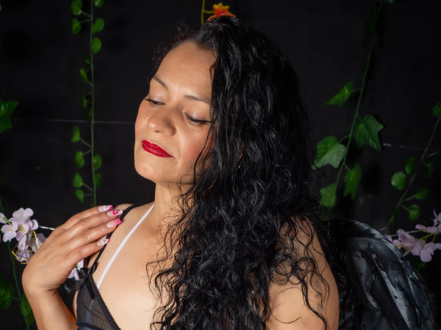Alexandra Jones mit lockigem, schwarzem Haar, rotem Lippenstift und schwarzen Flügeln, mit geschlossenen Augen, die ihre Brust berührend, mit rosafarbenen Nägeln, vor einem dunklen Hintergrund mit grünen Blättern und rosa Blumen..