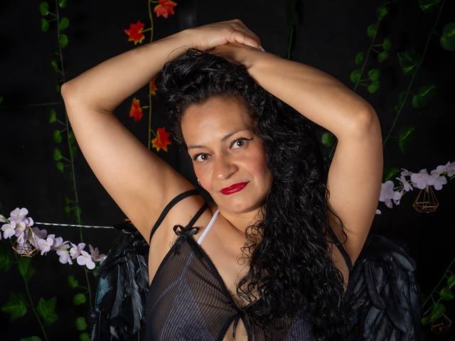 Alexandra Jones, con i capelli neri ricci, le braccia alzate, indossa lingerie nera trasparente, rossetto rosso, sorride. Sullo sfondo nero ci sono foglie verdi e fiori rosa. Il seno è visibile attraverso il tessuto trasparente..