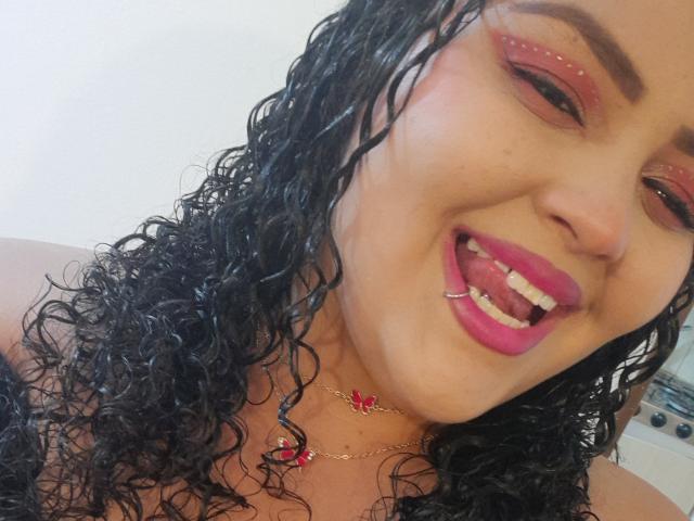 MelannieCarter has curly black wet hair, pink lipstick, pierced tongue, red eye makeup, gold necklace with red gem, smiling, showing teeth, with a light brown skin tone.
