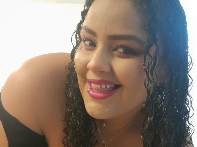 MelannieCarter with curly black hair, brown eyes, pink lipstick, smiling, wearing a black top, showing her shoulder and upper chest, looking cute and sexy.