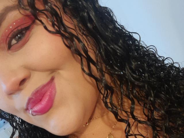 MelannieCarter's face with curly black hair, pink lipstick, pierced lower lip, red eyeshadow, and gold necklace, smiling slightly, showing her cute nose and smooth brown skin.