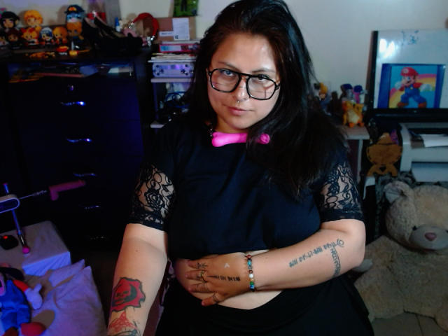 LilithHeavy with glasses, black lace shirt, pink bowtie, black hair, tattoos on arms, holding belly, sitting in room, surrounded by toys and stuffed animals.