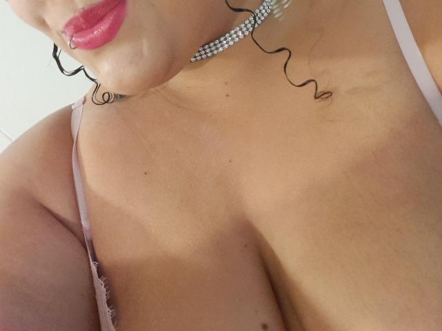 MelannieCarter's chest with tan lines, wearing pink lace bra, silver choker, red lipstick, and black curly hair, showing her plump tits and a small lip piercing.