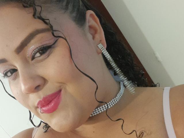 MelannieCarter has curly black hair, pink lipstick, and is wearing glittery eyeshadow, a diamond necklace, and matching earrings. Her tits are partially visible in a pink top.