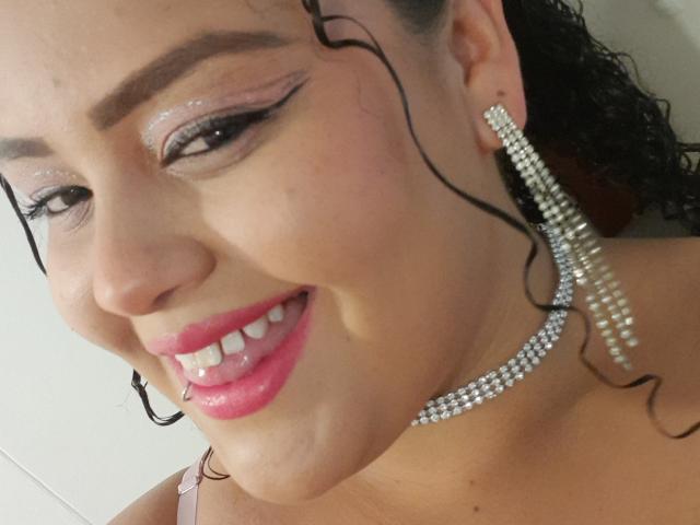 MelannieCarter's face with curly black hair, glossy pink lipstick, shiny eyeshadow, and large diamond earrings. Her smile shows white teeth. She's wearing a pink strap.