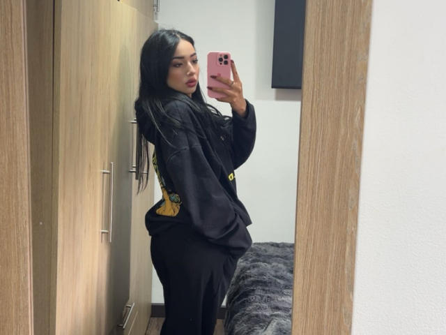 LillyTaylor with long black hair, wearing a black shirt and black pants, taking a mirror selfie, showing her ass, holding a pink phone, in a wooden room.