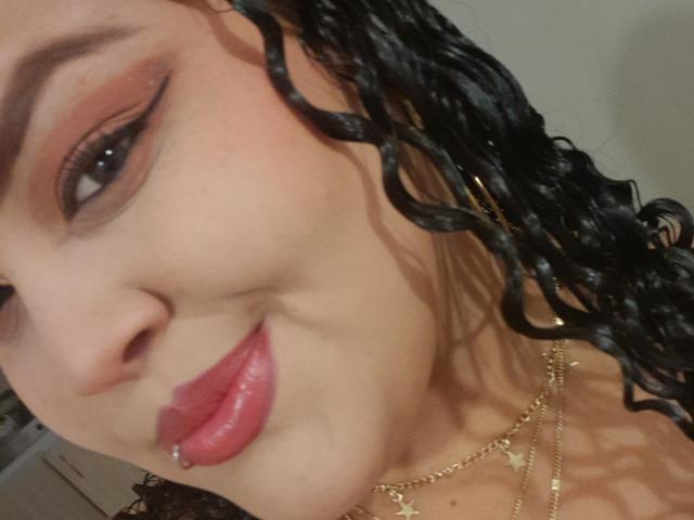 MelannieCarter's face, showing curly black hair, red lipstick, and a gold necklace. Her eye has smoky makeup, and she has a lip piercing. Her skin is light brown.