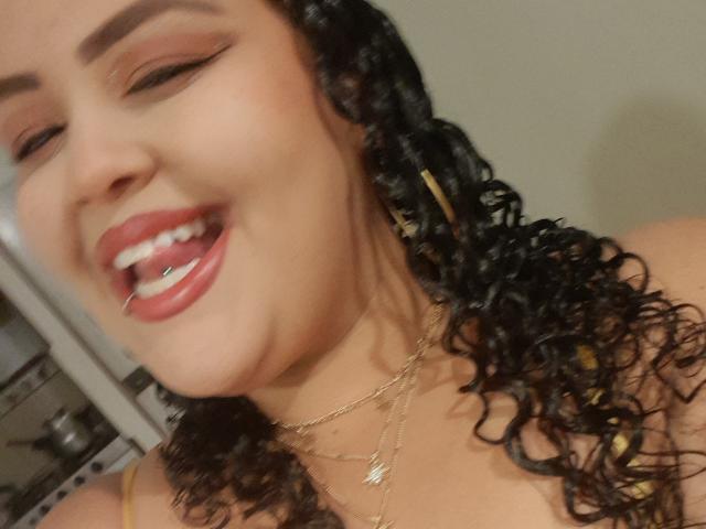 MelannieCarter with curly black hair, gold earrings, and a gold necklace, smiling with her mouth open, showing a silver tooth. Her eyes are closed, and she has red lipstick.