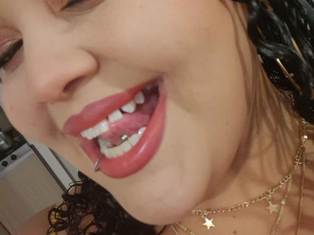 MelannieCarter's face showing a wide open mouth with red lipstick, a tongue piercing, and white teeth. Her skin is tan, and she's wearing a gold necklace with star charms.