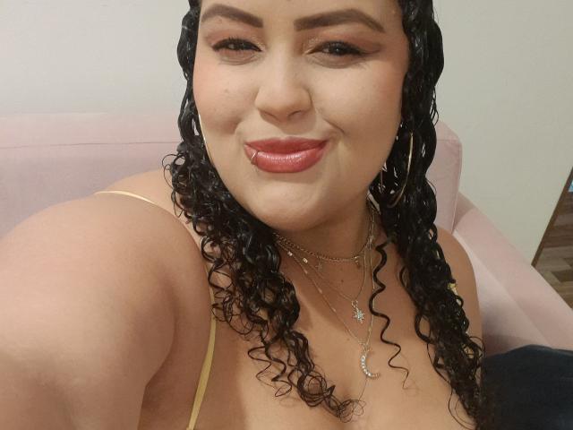 MelannieCarter has curly black hair, hoop earrings, and red lipstick, smiling with her tongue slightly out. She's wearing a yellow top and multiple necklaces, showing her tits.