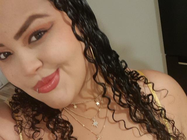 MelannieCarter with curly black hair, red lipstick, and a lip piercing, wearing gold necklaces, smiling at the camera, showing her tits in a yellow top.