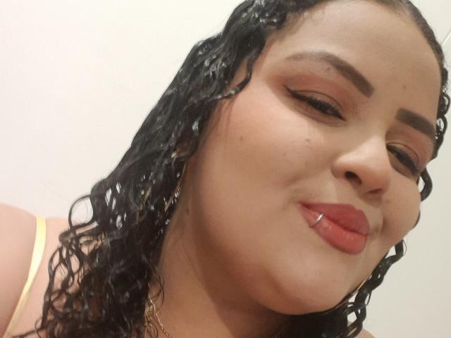 MelannieCarter has wet, curly black hair, full lips with red lipstick, and brown eyes with thick eyebrows, wearing a yellow top, smiling with a slight pout.