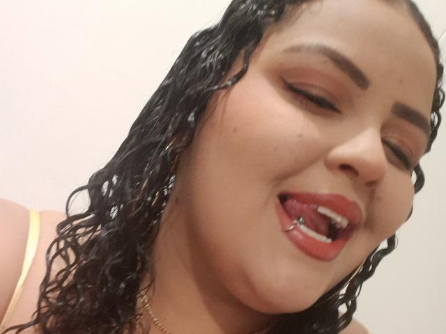 MelannieCarter has wet, curly black hair, brown eyes, and a tongue piercing with a small ball, wearing red lipstick and a yellow top, smiling with eyes closed.