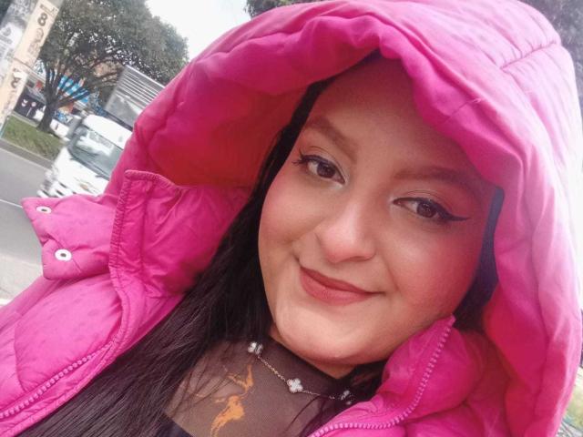 VictoriaRiver with pink puffy jacket hood, black eyeliner, small smile, black hair, light skin, necklace with small beads, tattoo on chest, standing outside, car in background.