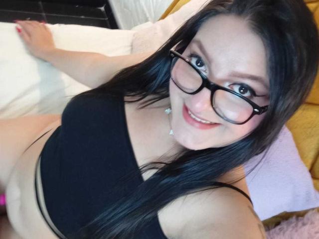 VictoriaRiver, with black glasses and long black hair, smiling at the camera, wearing a black sports bra, showing her tits and a hint of her pink pussy.