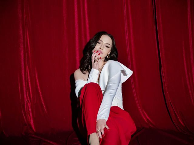 ElliMorgan with wavy dark hair, white off-shoulder top, and red pants, sits against a red velvet background. She's slightly hunched, right knee up, left hand touching her face with red-painted nails. White top has ruffled sleeves. Red velvet background creates a rich, textured contrast with her white top and red pants. Lighting highlights her face and hands. She looks relaxed and confident. The image is sharp, with a clear focus on ElliMorgan.
