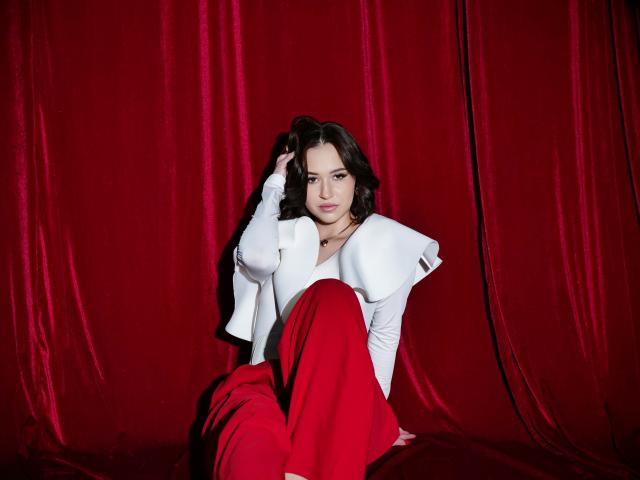 ElliMorgan with medium tits, white puffy-sleeve top, and red pants, sitting against red velvet, hand in dark wavy hair, slight smile, confident, sexy.
