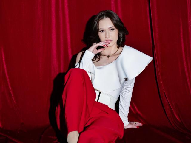 ElliMorgan with wavy black hair, wearing a white top with a flared collar and red pants, sitting against a red velvet backdrop, touching her pink-lipsticked mouth.