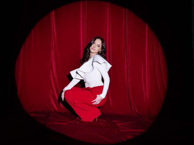 ElliMorgan with long dark hair, white top, and red pants, crouching on red velvet, wearing white gloves, looking at camera with a slight smile, in a circular frame.