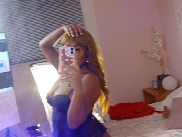 AmberQueeny mit langen blonden Haaren, Brille und einem schwarzen Spitzenkleid, die in einem Spiegel-Selfie ihre Brüste zeigt, wobei sie ihr Haar hochhält, in einem rosafarbenen Schlafzimmer..