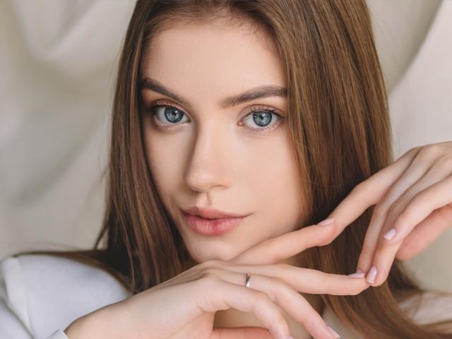 Close-up portrait of EricaDiaz. She has fair skin, straight brown hair, and striking blue eyes. Her eyebrows are neatly shaped, and she has a subtle, natural smile. She rests her fingers gently on her chin, showing a thin, silver ring on her right index finger. Her nails are neatly trimmed. The lighting is soft, highlighting her clear complexion and smooth skin. The composition focuses on her face, with her hair framing her face on both sides. Her expression is calm and slightly curious.